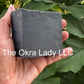 A hand holding a rectangular dark gray soap bar in front of green leaves, shown within a mobile app screenshot. The soap bar has white text reading 'The Okra Lady LLC' across the center.