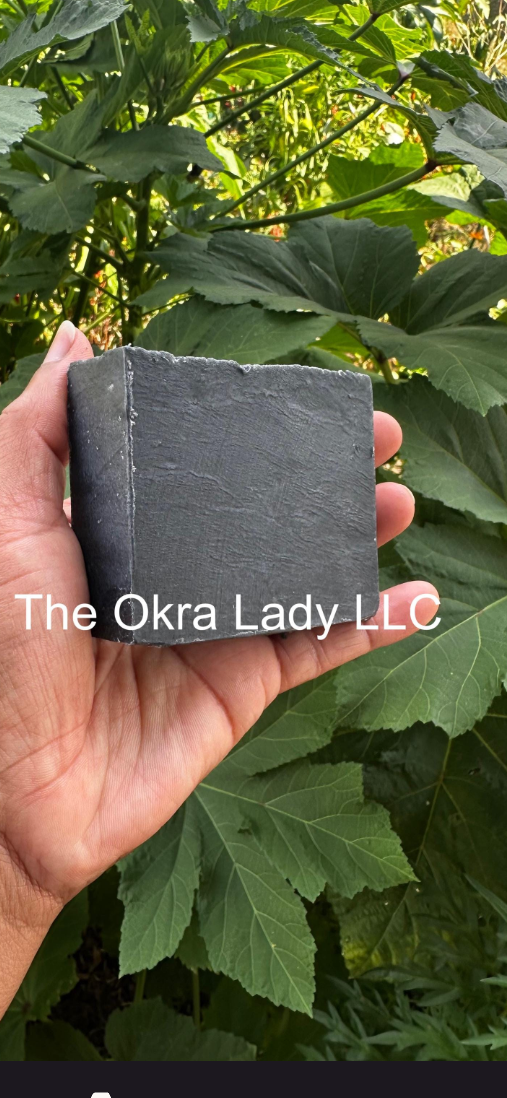 A hand holding a rectangular dark gray soap bar in front of green leaves, shown within a mobile app screenshot. The soap bar has white text reading 'The Okra Lady LLC' across the center.