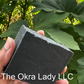 A hand holding a rectangular bar of solid black soap outdoors in front of green leaves. The soap has white text reading 'The Okra Lady LLC' across the center.