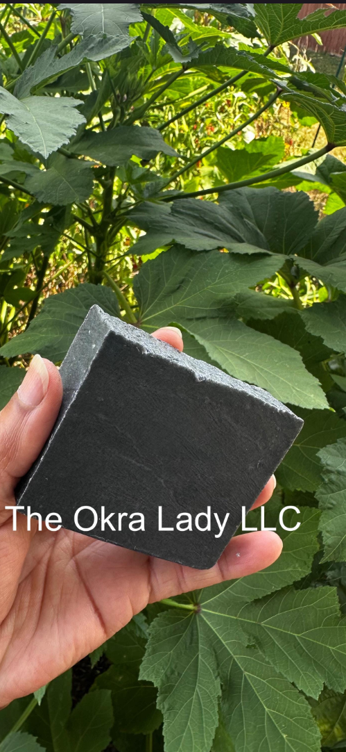 A hand holding a rectangular bar of solid black soap outdoors in front of green leaves. The soap has white text reading 'The Okra Lady LLC' across the center.